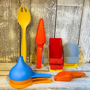 May include: A collection of colorful plastic kitchen utensils, including a yellow fork, orange citrus reamer, red container, blue and orange funnel, and orange scoop. The items are arranged against a weathered wood background.