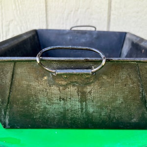 May include: A rectangular, galvanized metal planter with a dark, weathered finish. It features two arched handles and a tapered design. The planter is sitting on a green surface.