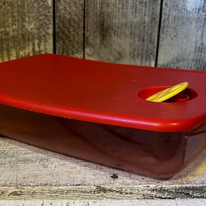 May include: A rectangular, red plastic food storage container with a matching lid. The lid has a small, circular opening with a yellow and brown utensil. The container is on a white wooden surface.