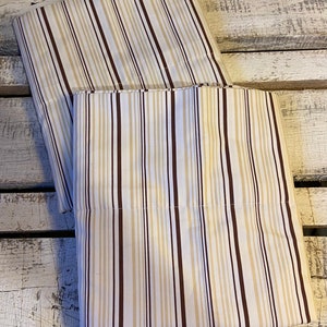 May include: Two folded pillowcases with a vertical striped pattern. The stripes alternate between white, tan, and dark brown. The pillowcases are made of a soft fabric and are neatly folded.