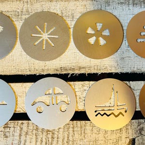 May include: Eight circular stencils with various cut-out designs, including a heart, snowflake, and car. The stencils are made of metal and are in shades of silver and gold. They are arranged on a rustic wooden surface.