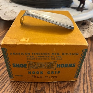 May include: A vintage shoe horn made of nickel-plated metal, resting on a yellowed cardboard box. The box features text that reads "SHOE BAKBONE HORNS" and "HOOK GRIP". The box is from American Findings Mfg. Division, Steel Industries, Inc.