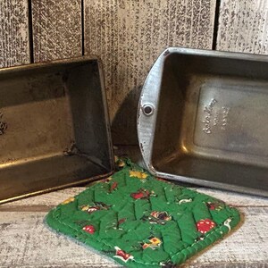 May include: Two rectangular metal loaf pans with rounded corners and a green quilted potholder featuring a small floral pattern. One pan is stamped with the text EKCOLOY Made in USA, while the other pan has a metallic finish and side handles.
