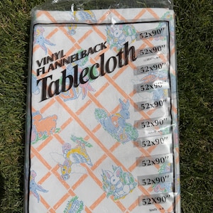 May include: A packaged vinyl flannelback tablecloth with a repeating pattern of pastel-colored bunnies, birds, and other figures on a white background with an orange lattice design. The tablecloth measures 52 x 90 inches.