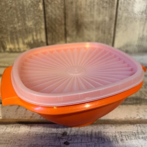 May include: An orange plastic bowl with a pink lid. The lid is square with a sunburst design and fits snugly on the bowl. The bowl has two orange handles on either side. This vintage Tupperware container is ideal for food storage.