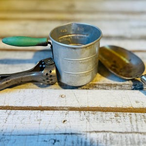 May include: A collection of vintage kitchen tools including a silver metal measuring cup with embossed markings, a pair of metal tongs with green wooden handles, and a metal scoop featuring a matching green wooden handle and a worn finish.