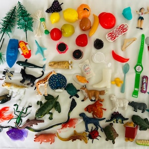 May include: A collection of colorful miniature toys and decorative items, including fruits, animals, and accessories. The assortment features a variety of shapes and sizes, with a mix of plastic and other materials. The toys are arranged on a white surface.