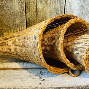 May include: A set of three nested cornucopia baskets crafted from light brown woven wicker. Each basket features a tapered, horn-like shape with a wide, circular opening finished with a braided rim. The baskets are stacked horizontally.