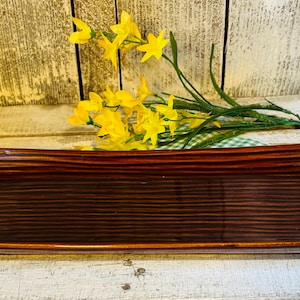 May include: A rectangular, brown ceramic serving platter with a textured, ridged surface. The platter is displayed on a white wooden surface, with yellow flowers and greenery in the background.