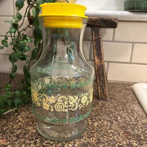 May include: A clear glass pitcher with a bright yellow plastic lid. The body of the pitcher features a decorative band of yellow and green floral and scroll patterns encircling the center. The glass has a wide base and a tapered neck design.