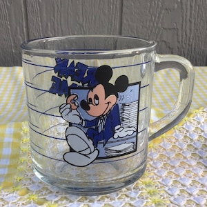 May include: Clear glass mug featuring Mickey Mouse in a suit, holding a stack of papers. The mug has blue text and lines, with a handle on the side. The background is a yellow and white checkered pattern.