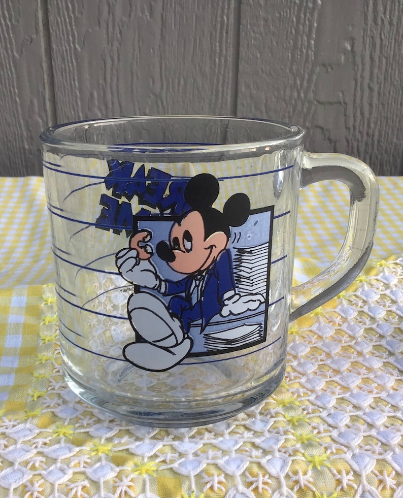 May include: Clear glass mug featuring Mickey Mouse in a suit, holding a stack of papers. The mug has blue text and lines, with a handle on the side. The background is a yellow and white checkered pattern.