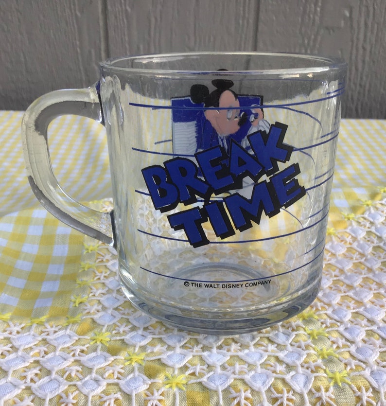 May include: Clear glass mug with a handle featuring a graphic of Mickey Mouse and the words "BREAK TIME" in blue. The mug has blue horizontal lines and the text "THE WALT DISNEY COMPANY" at the bottom.