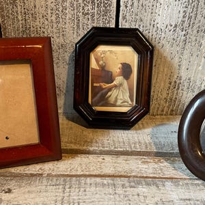 May include: Three picture frames: a rectangular reddish-brown frame, an octagonal dark brown frame containing a print of a person playing a piano, and a smooth, dark brown oval frame. Each frame is distinct in shape and color, showcasing varied designs.