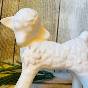 May include: A white ceramic lamb figurine featuring a textured, curly wool surface and a small blue painted detail on the neck area. The lamb is depicted in a standing position with its head turned slightly to the side and ears pointed outward.