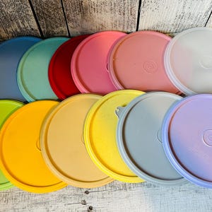 May include: A collection of colorful, round plastic lids in various shades, including green, yellow, pink, and blue. The lids are arranged in a row, showcasing their diverse hues and simple design. They appear to be for food storage containers.