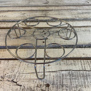 May include: A circular silver-toned metal wire rack featuring a central ring surrounded by six smaller evenly spaced rings. The structure is supported by three vertical wire legs that extend downward from the outer frame to provide elevation.