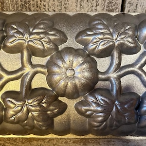 May include: A rectangular metallic baking pan featuring a raised, embossed design of pumpkins and leaves across the entire surface. The pan has a silver-toned finish with a textured appearance and integrated handles on the left and right sides.