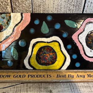 May include: Rectangular black decorative tile with colorful abstract design. Features pink, yellow, white, and blue shapes. The tile has a glossy finish and is mounted with metal hardware. The text "MEADOW GOLD PRODUCTS - Best By Any Measure" is visible.