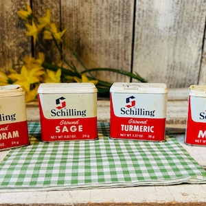May include: Four vintage rectangular metal spice tins by Schilling, featuring white tops and red bases with blue logos. The labels read Ground Marjoram, Ground Sage, Ground Turmeric, and Ground Mace, with net weights of 0.87 oz, 1.12 oz, and 38 g.