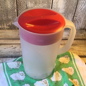 Pu&ograve; includere: Una caraffa in plastica traslucida con coperchio rosso e manico, dotata di un'indicazione di capacit&agrave; di 1 gallone sul lato. La caraffa &egrave; posizionata su un panno di tessuto bianco decorato con un motivo ripetitivo di piccole pecore bianche.