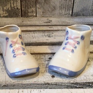 May include: A pair of cream-colored ceramic baby shoes featuring hand-painted details. Each shoe has light pink laces, blue eyelets, and a blue toe cap. The glossy finish covers the entire surface of these decorative ceramic footwear items.