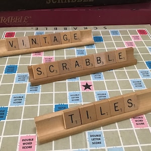 May include: A close-up of a Scrabble game board with three wooden racks holding the tiles spelling out "VINTAGE", "SCRABBLE", and "TILES".