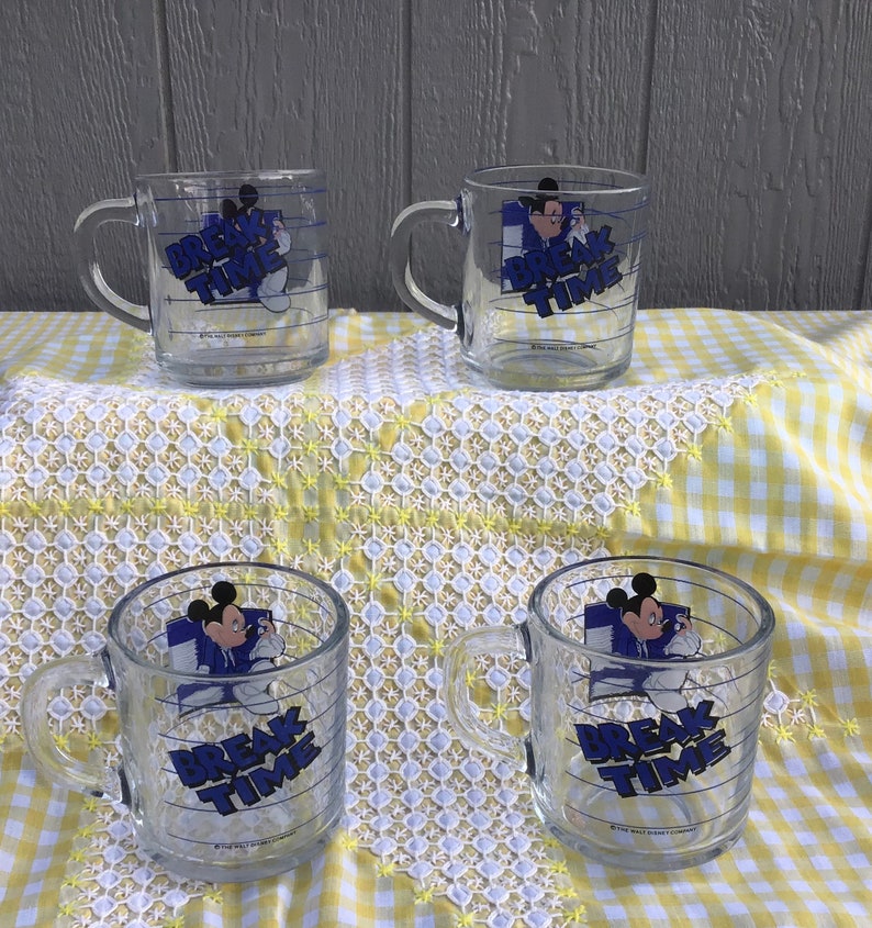 May include: Four clear glass mugs with handles, each featuring a graphic of Mickey Mouse and the words "BREAK TIME" in blue. The mugs are on a yellow and white checkered tablecloth.