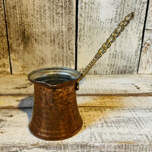 May include: A copper-colored Turkish coffee pot, also known as a cezve, with a long, ornate handle. The pot has a wide base that tapers to a narrower top. The handle is gold-toned with a decorative pattern. The pot is designed for brewing coffee.