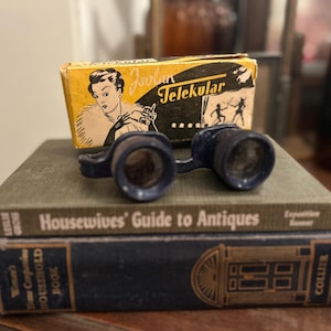 May include: A pair of small, dark blue binoculars sits atop a stack of vintage books. A yellow box with the word "Telekular" and an illustration of a woman is behind the binoculars. The books are titled "Housewives' Guide to Antiques" and "Women's Home Companion Household Book."