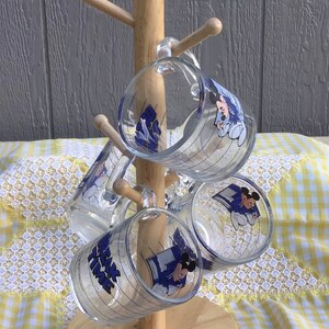 May include: A wooden mug tree holds six clear glass mugs. Each mug features a blue and white graphic of Mickey Mouse and the text "Mickey's Bake Time". The mugs are arranged on a light wood stand against a gray background.