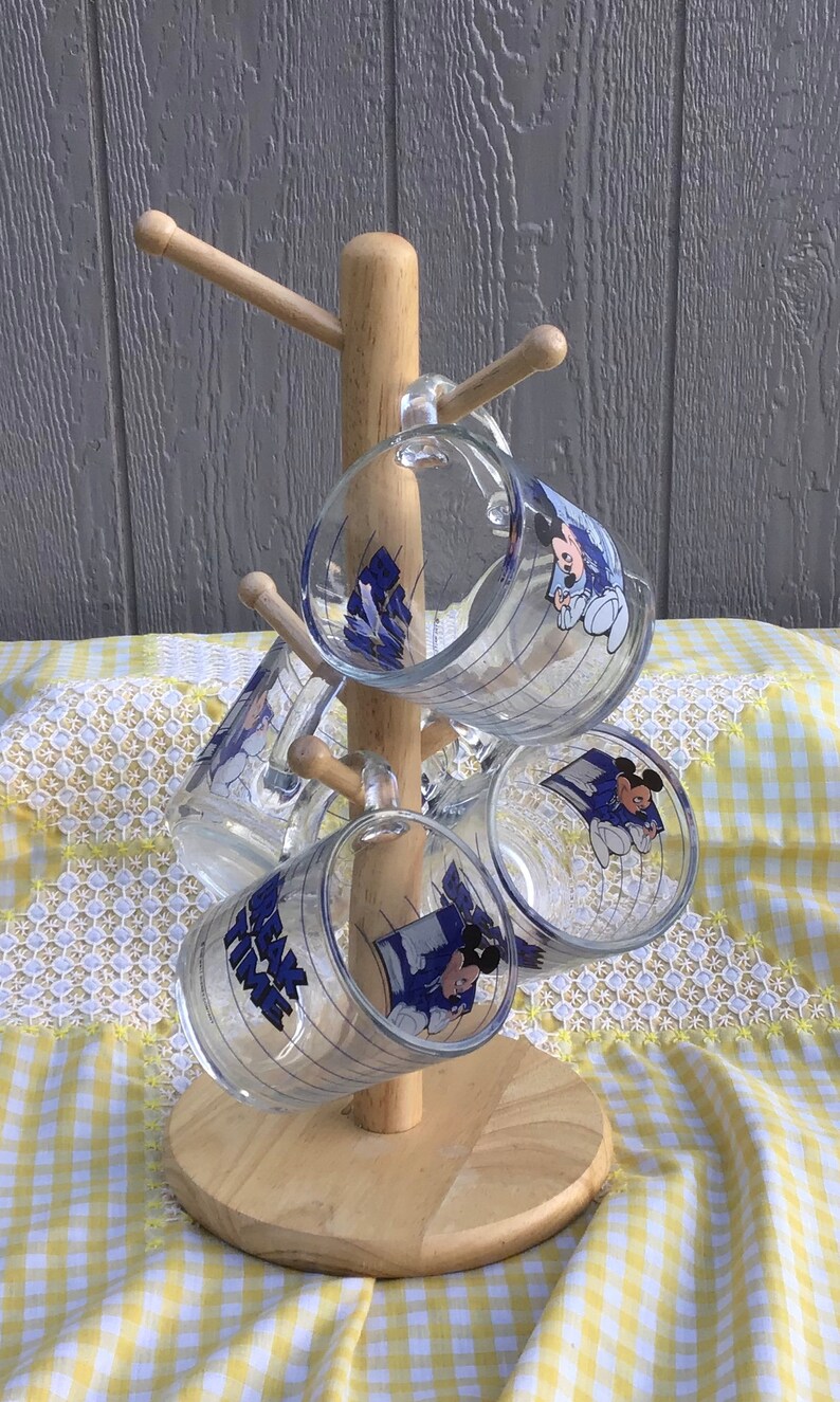 May include: A wooden mug tree holds six clear glass mugs. Each mug features a blue and white graphic of Mickey Mouse and the text "Mickey's Bake Time". The mugs are arranged on a light wood stand against a gray background.
