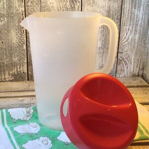 Pu&ograve; includere: Una caraffa in plastica bianca traslucida con manico e beccuccio, accompagnata da un coperchio rotondo rosso separato dotato di due aree di presa incassate e una piccola apertura circolare sul lato per versare i liquidi.