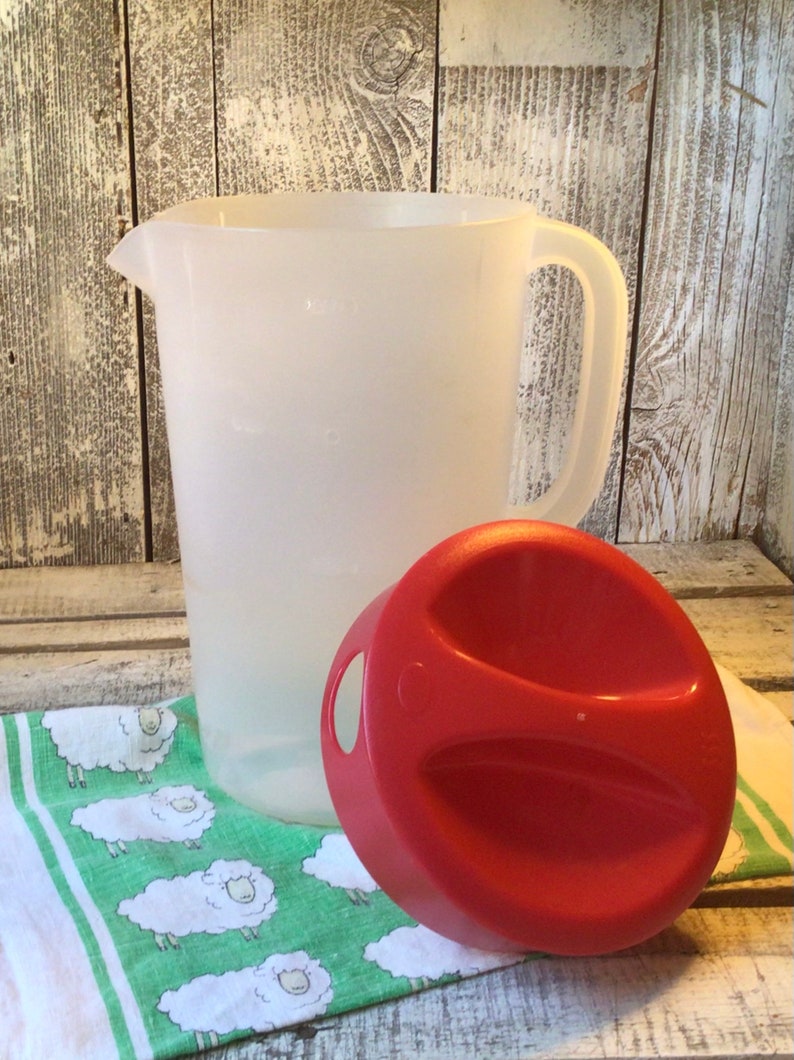 Pu&ograve; includere: Una caraffa in plastica bianca traslucida con manico e beccuccio, accompagnata da un coperchio rotondo rosso separato dotato di due aree di presa incassate e una piccola apertura circolare sul lato per versare i liquidi.