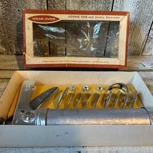 May include: A silver metal Wear-Ever cookie gun and pastry decorator set in a rectangular box. The set includes a cylindrical tool with a trigger handle and several metal discs for shaping dough. The box lid features the text Wear-Ever.