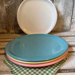 May include: A stack of three round plates in light blue, yellow, and pink, with a single white plate positioned upright behind them. The plates have smooth, solid-colored surfaces and raised edges, resting on a green and white checkered napkin.
