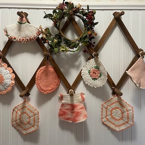 May include: A collection of handmade crochet items in shades of white and peach, including pot holders and a decorative dress shape, displayed on a wooden rack. A small floral wreath is at the top. The items are hanging on a wooden rack against a white wall.