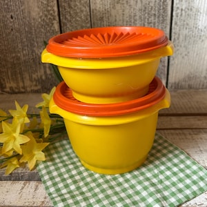 May include: Two yellow plastic bowls with orange lids are stacked on a green and white checkered cloth. The lids have a ridged design. The bowls are on a wooden surface with yellow flowers in the background. Kitchenware.