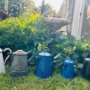 May include: A collection of vintage enamelware coffee pots in various colors, including white, gray, and blue, with black accents. The pots are arranged on a grassy lawn, with a backdrop of lush greenery and a yellow and white chair.