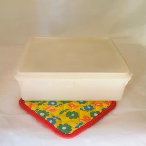 May include: A square, off-white plastic container with a matching lid sits atop a yellow, floral-patterned trivet with a red border. The trivet features blue, orange, and green flowers.
