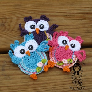 May include: Three crocheted owls, one purple, one blue, and one pink, with white eyes and orange beaks. Each owl has a small flower on its wing. The owls are on a wooden surface. The text "Made with Love" is visible in the image.