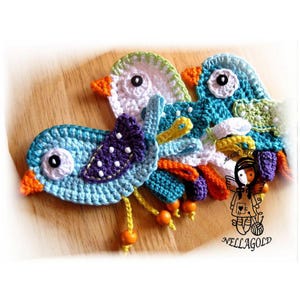Könnte beinhalten: Drei gehäkelte Vögel mit bunten Akzenten und Holzperlen. Die Vögel sind blau, weiß und grün mit violetten und orangen Details.