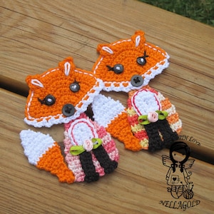 Può includere: Due ornamenti di volpe arancione all'uncinetto con bordino bianco, occhi neri e bottoni neri. Ogni volpe indossa un vestito a righe rosa, arancio e nero con un nastro verde e un fiore rosa.