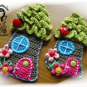 Könnte beinhalten: Zwei gehäkelte Häuser mit grünen Dächern, rosafarbenen Türen und blauen Fenstern. Jedes Haus hat einen Marienkäfer auf dem Dach und Blumen um den Sockel.