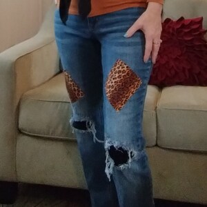 May include: A pair of blue jeans with distressed details and patches of leopard print fabric. The jeans are worn with a brown sweater and a brown belt.