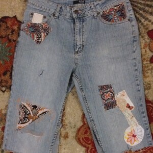 May include: Light blue denim shorts with patchwork details. The shorts have a frayed hem and are decorated with various fabric scraps in floral and paisley patterns. The brand name "ana" is visible on the waistband.