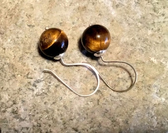 Puede incluir: Un par de pendientes con cuentas de piedra preciosa de ojo de tigre pulidas. Cada cuenta es una esfera de tonos marrón y dorado, montada sobre un gancho plateado. Los pendientes se exhiben sobre una superficie clara y moteada.