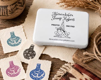 May include: A rectangular white ink pad labeled Abracadabra Stamp Makers with a frog wearing a wizard hat illustration. Four small square pieces of textured paper are displayed nearby, each featuring a stamped image of a vase in blue, black, or purple ink.