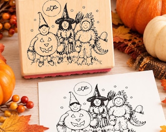 May include: A rectangular wooden rubber stamp featuring a black ink illustration of three children in Halloween costumes: a pumpkin, a witch with a broom, and a dinosaur. A small bat is depicted in a circle above them, surrounded by stars.