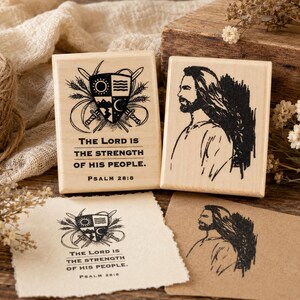 May include: Two rectangular wooden stamps with black ink designs. One stamp features a shield with a sun, waves, mountains, and a crescent moon, with the text "THE LORD IS THE STRENGTH OF HIS PEOPLE. PSALM 28:8". The other shows a portrait of a man.